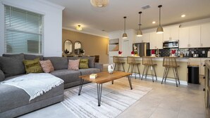 A modern living room with a grey sofa, a wooden coffee table, and a kitchen area with white cabinets and a black trash bin. - WonderFull 4Bd with Pool at Champions Gate 1174 (Davenport)