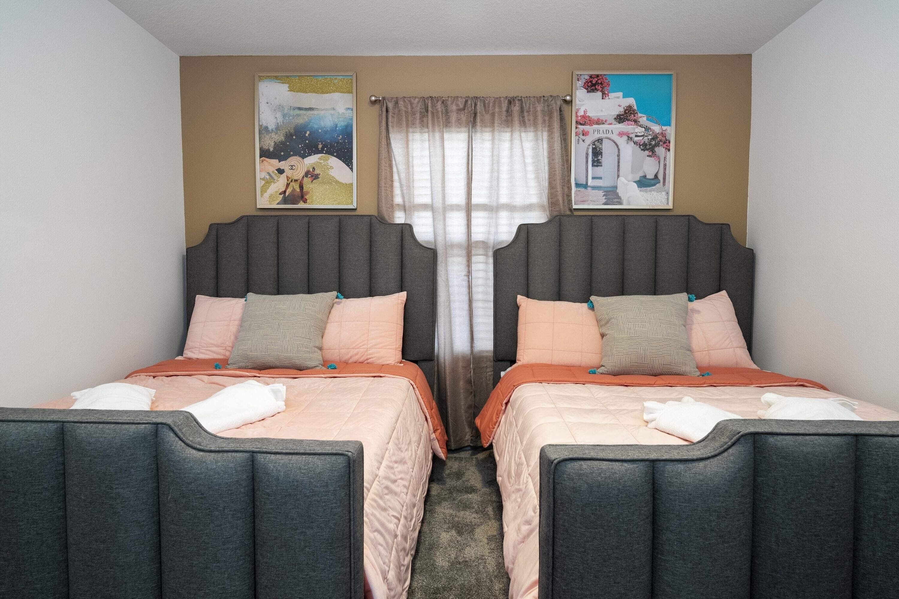 Een tweepersoonskamer met twee bedden, elk met een grijze hoofdbord en perzikkleurig beddengoed. Er is een raam met dunne gordijnen en twee ingelijste foto's aan de muur.