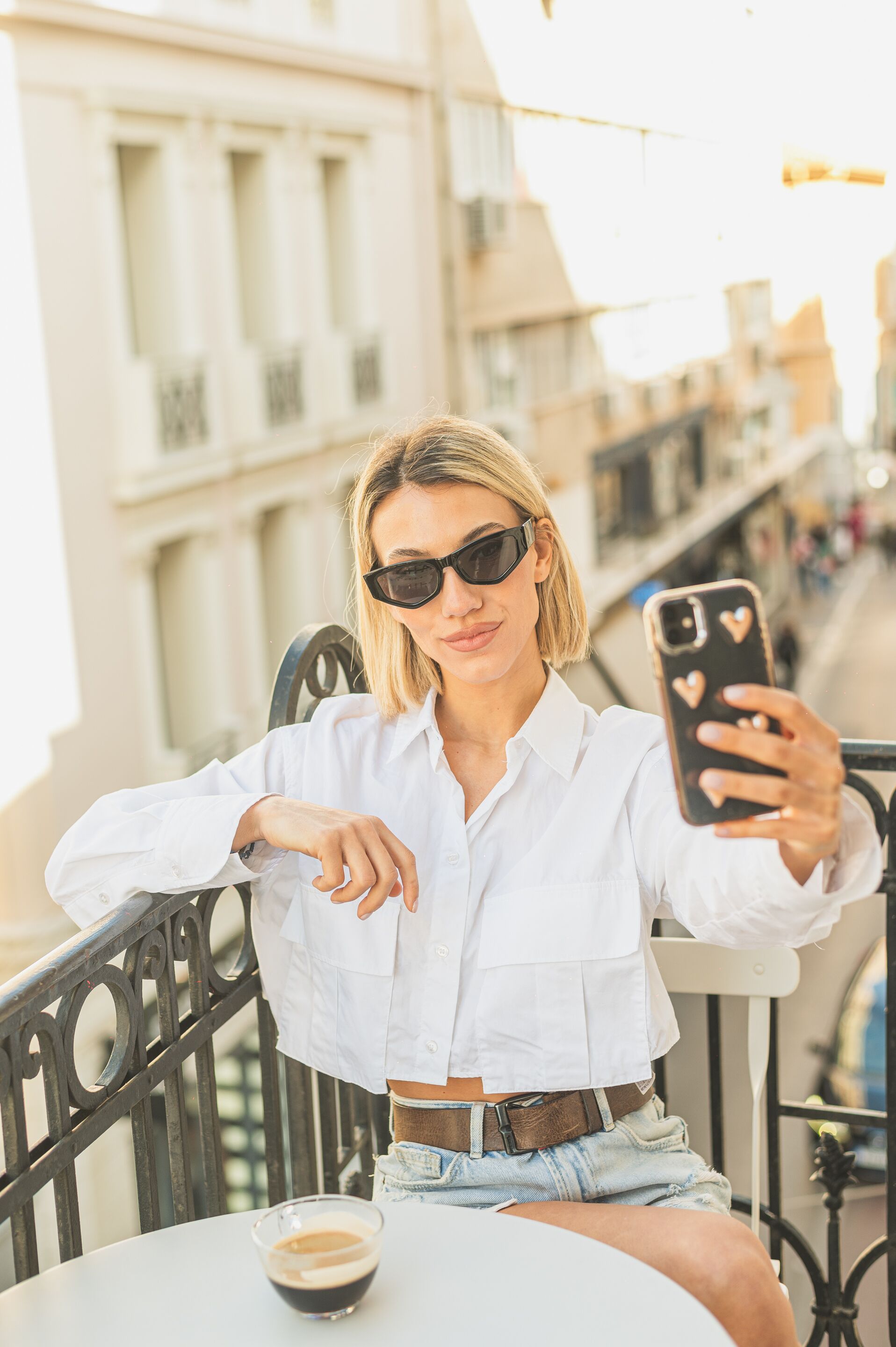 A person seated at a table with a smartphone, a cup of coffee, and a metal railing in the background.