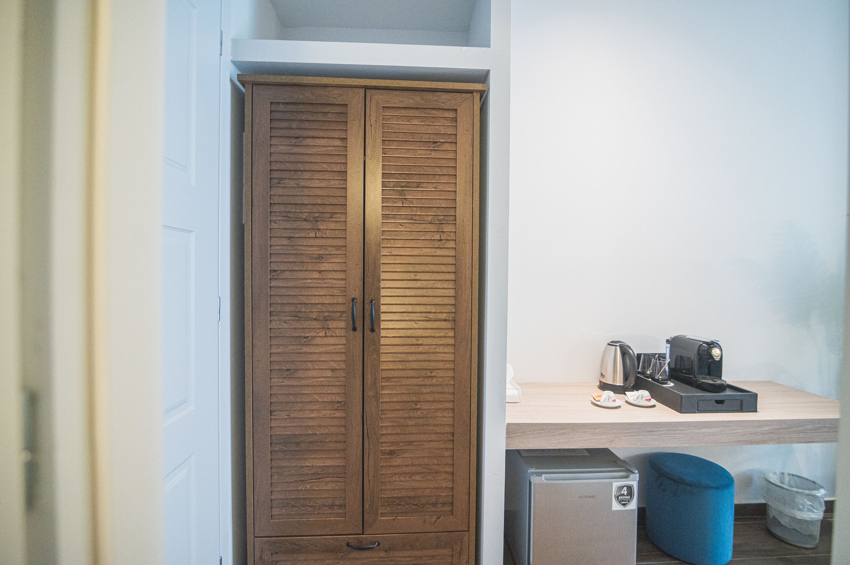 A compact room with a wooden wardrobe, a small kitchenette with a coffee maker, and a trash bin.
