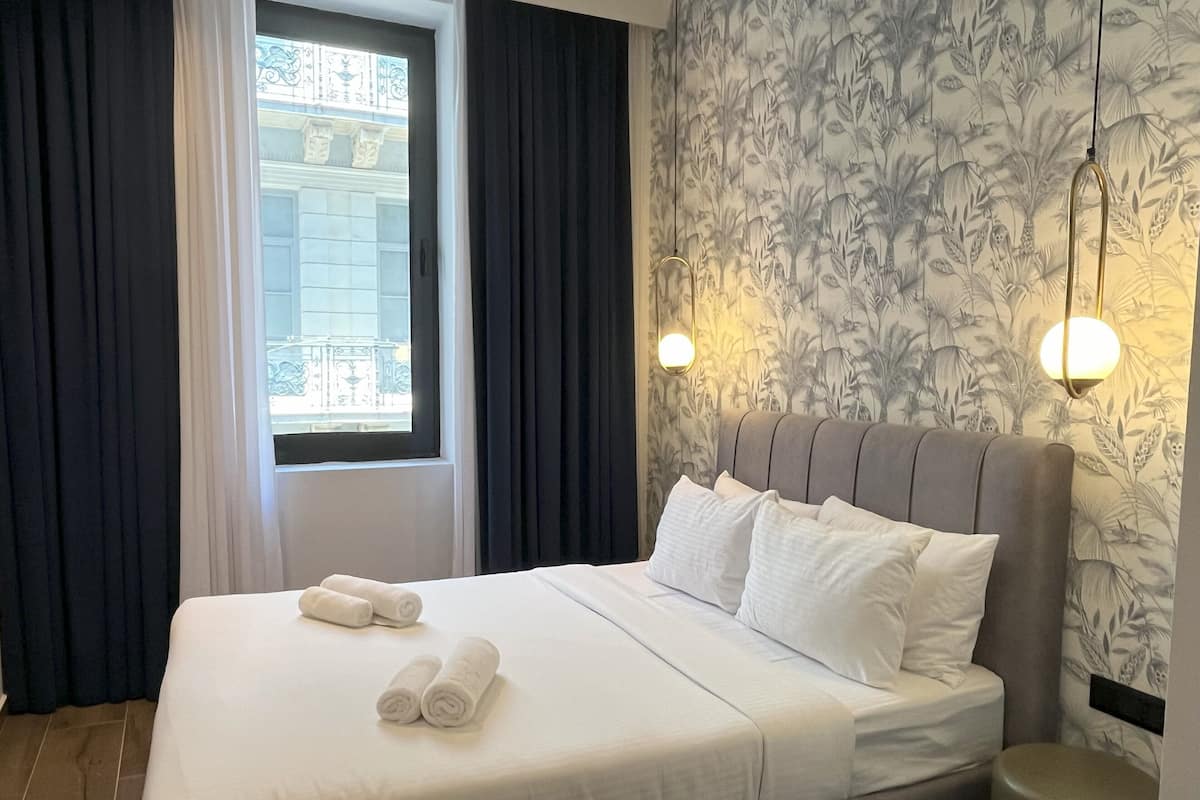 A neatly made bed with white linens and two rolled towels, a window with curtains, and a wall with a floral wallpaper pattern.