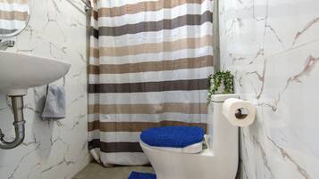 Un baño con cortina de ducha a rayas, un inodoro con tapete azul, un lavamanos y una pequeña planta en la pared.
