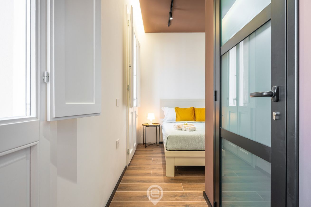 Un couloir étroit mène à une chambre à coucher qui contient un lit, une table de chevet et une lampe.
