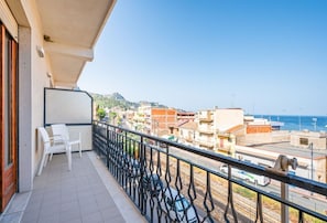 Un balcon donnant sur un paysage urbain côtier, une chaise blanche et une rambarde noire.