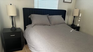 Un lit bien fait, recouvert d’une couvre-lit grise et agrémenté d’oreillers, flanqué de deux tables de chevet noires avec des lampes, dans une pièce dont le sol est carrelé et qui possède une tête de lit sombre.