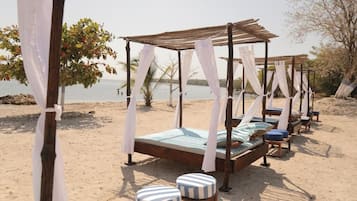Relax in a serene beach setting with elevated beds under canopies, surrounded by lush trees and a calm body of water.