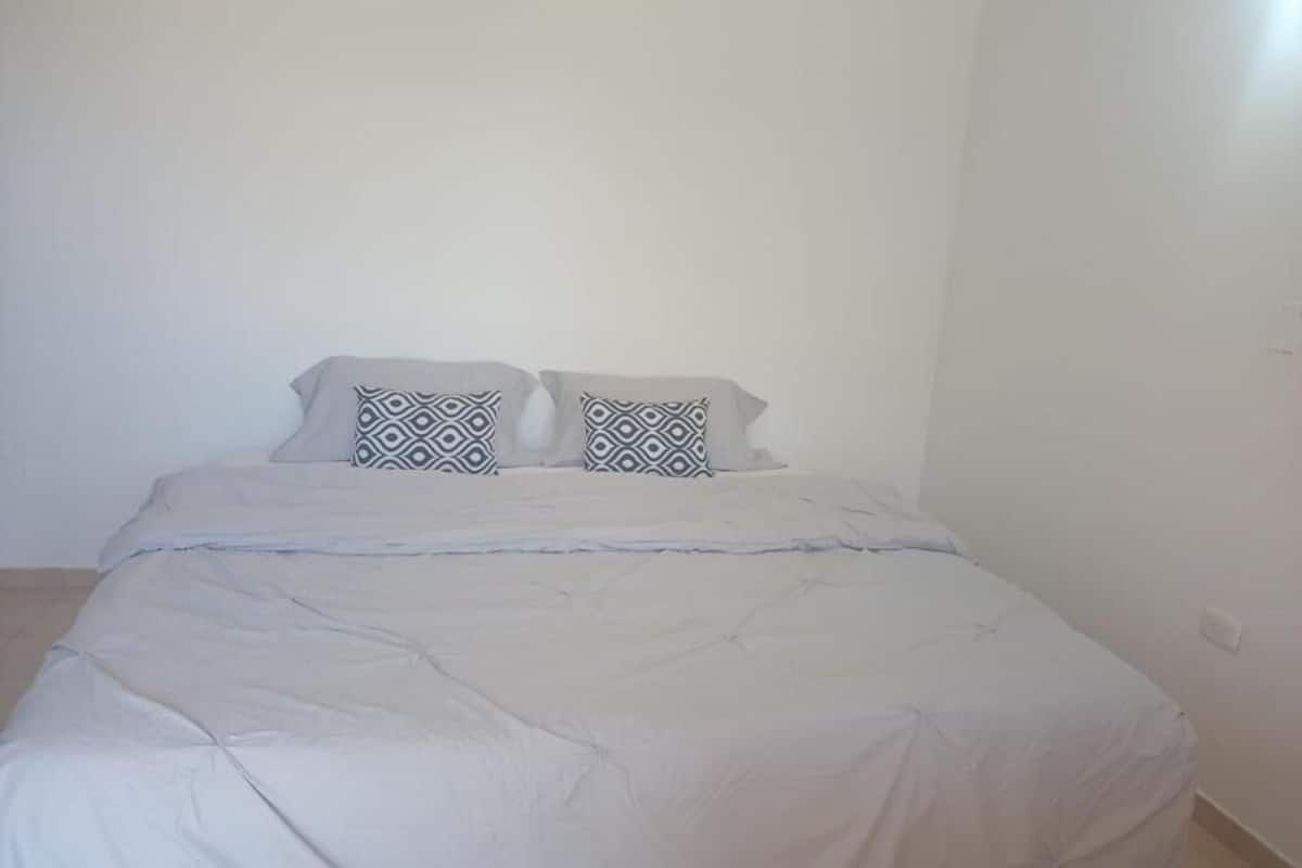 Una cama con edredón blanco y dos almohadas estampadas, una pared blanca lisa y un enchufe visible.