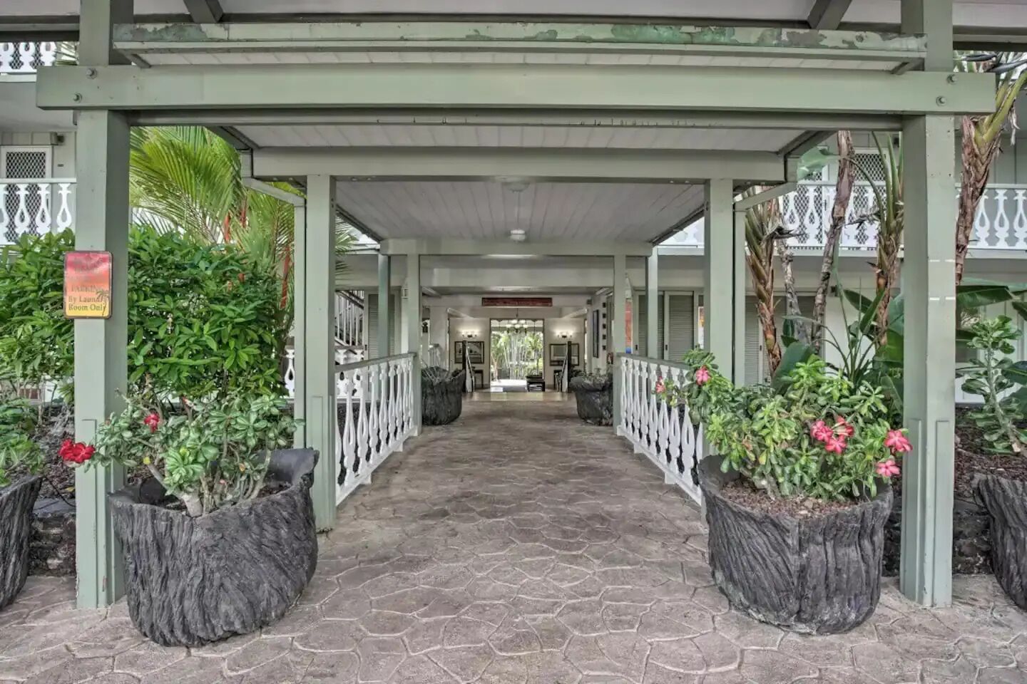 A covered walkway with stone flooring, flanked by planters with tropical plants and flowers, leading to a building entrance.