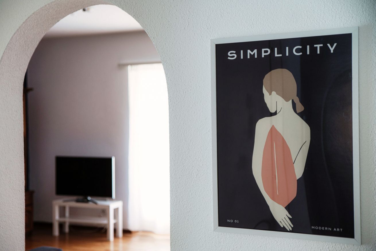 Une œuvre d’art encadrée, intitulée « SIMPLICITÉ », réalisée dans un style d’art moderne, représentant une figure minimaliste, avec une télévision et une petite table en arrière-plan.