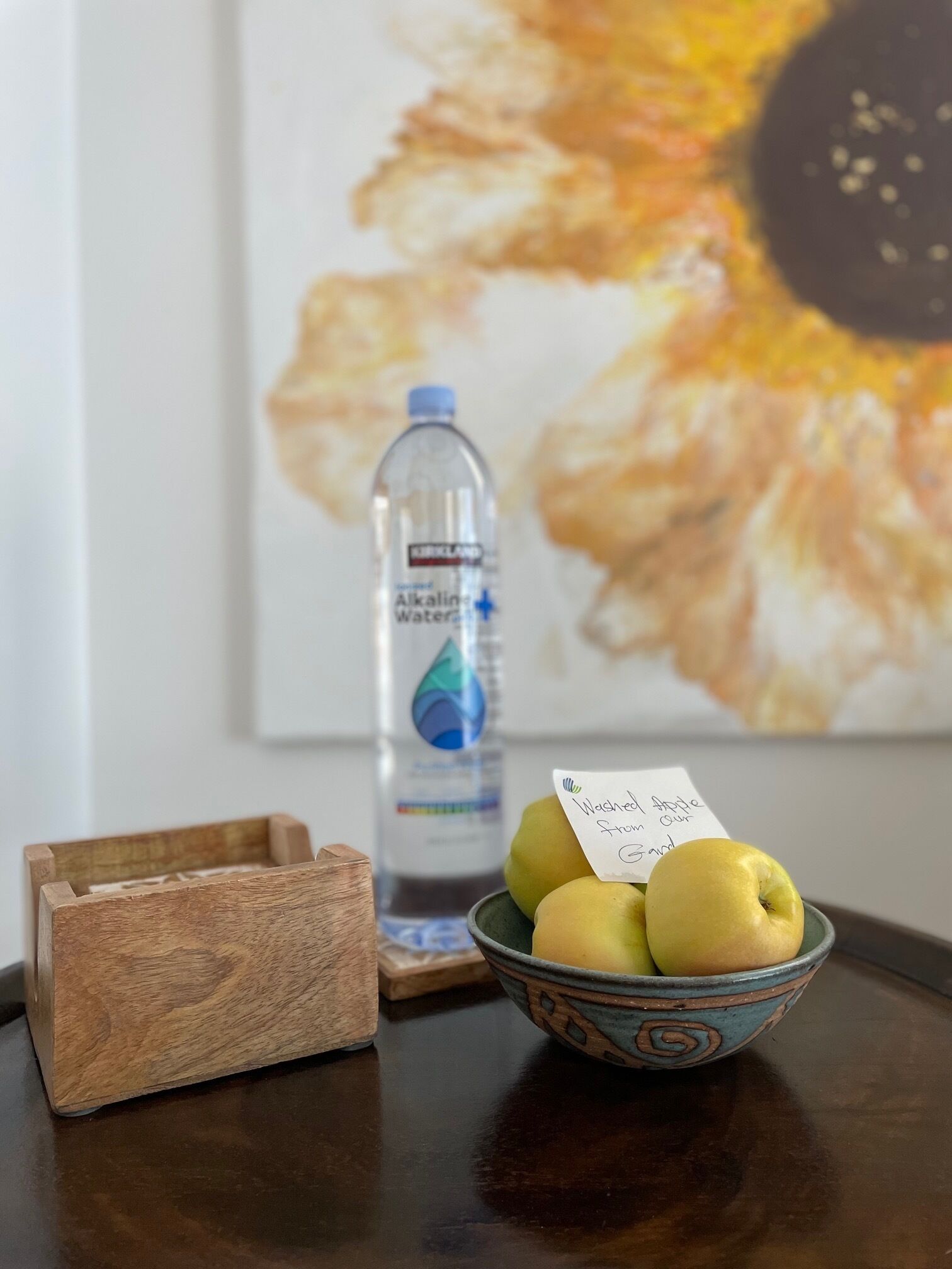 Una bandeja de madera, un tazón de manzanas y una botella de agua sobre una mesa con un cuadro enmarcado al fondo.