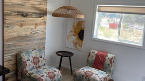 A modern living room with a wooden accent wall, two floral armchairs, a small round table, a hanging light fixture, a colorful rug, and a window with a view of greenery. - Wine Country Trail, View, Gated property, Detached Studio & Private View Patio, (Paso Robles)