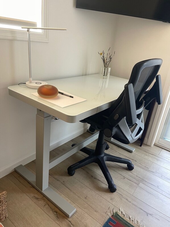 A modern office desk with a white surface, a black office chair, a small vase with dried plants, and a lamp.