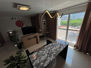 Una sala de estar moderna con televisor, mueble de entretenimiento de madera, mesa de comedor de mármol y balcón con vistas a edificios y vegetación.