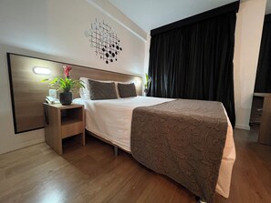 Una habitación de hotel moderna con una cama grande, una mesita de noche con lámpara, una planta en maceta y una obra de arte colgada en la pared.