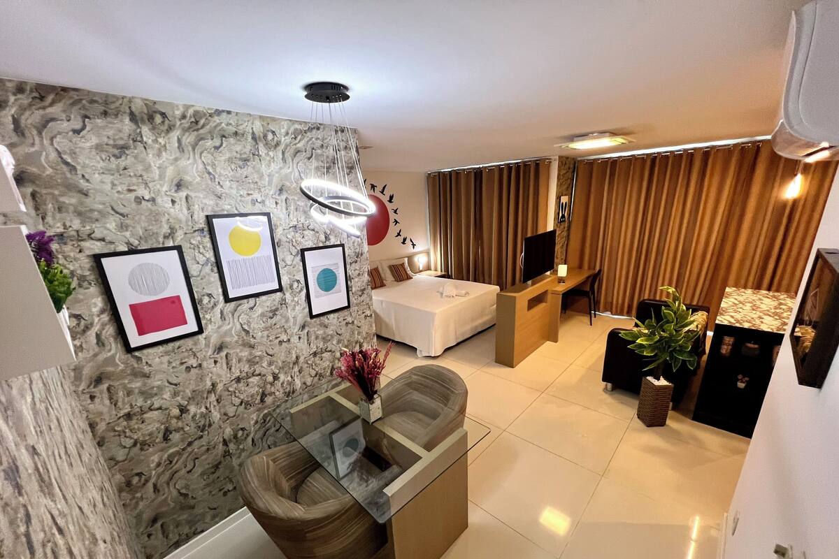 Habitación de hotel moderna con una pared de acento de mármol, una mesa de centro de cristal, un sofá, un escritorio con televisor y una planta en maceta.