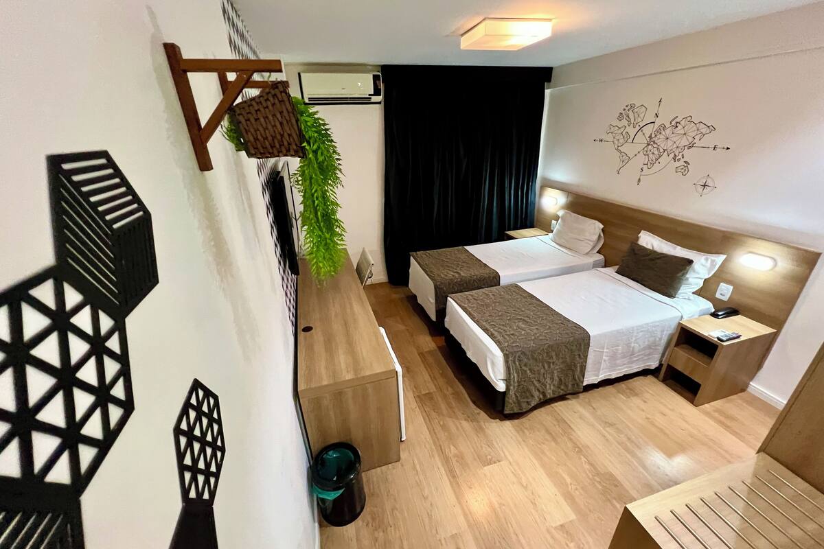 Habitación de hotel con dos camas, suelo de madera, aire acondicionado de pared, cortina negra, mapa en la pared y una mesita con lámpara.