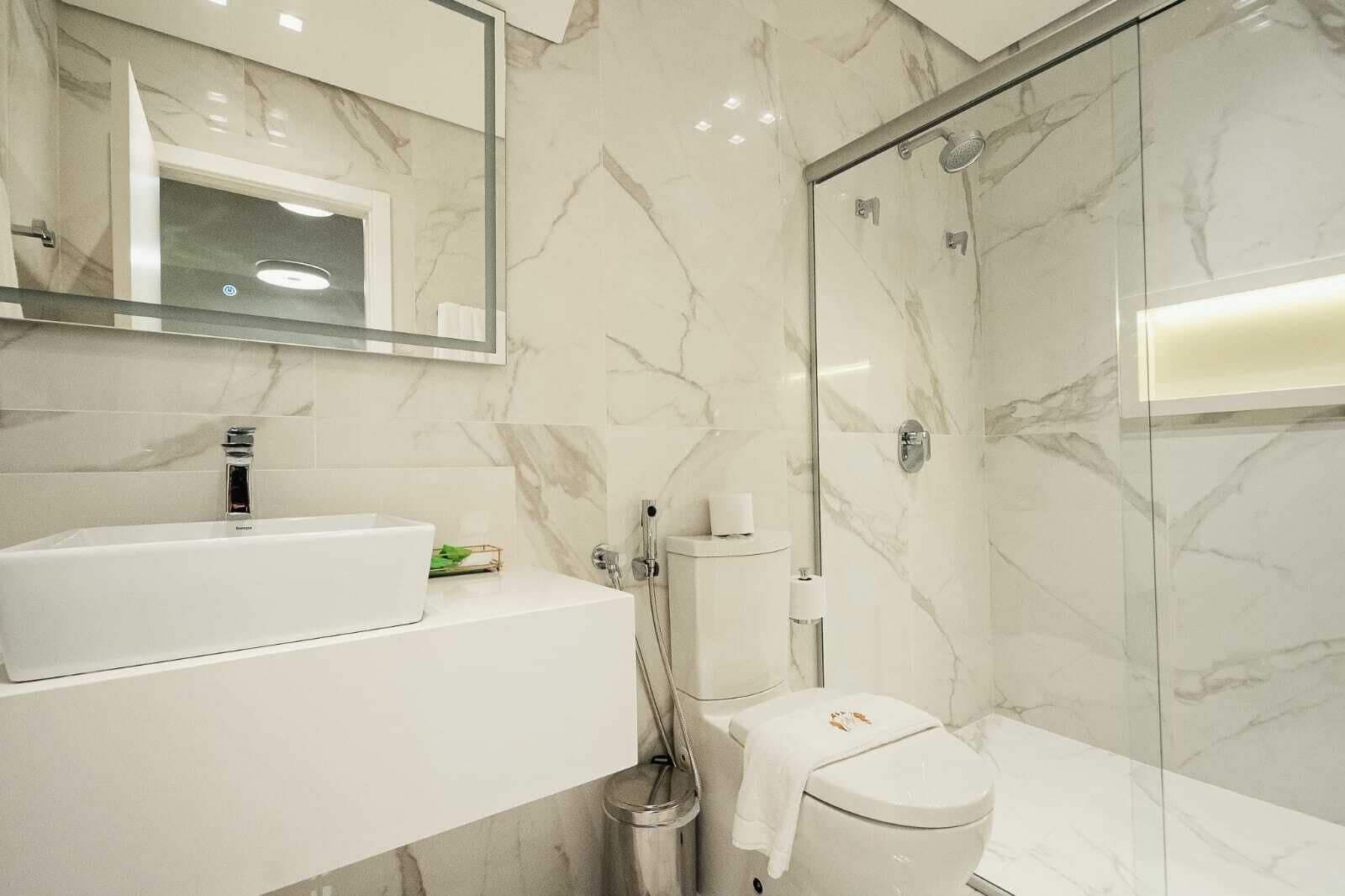 Un baño moderno con salpicadero de mármol, lavamanos blanco, inodoro y ducha con mampara de vidrio.