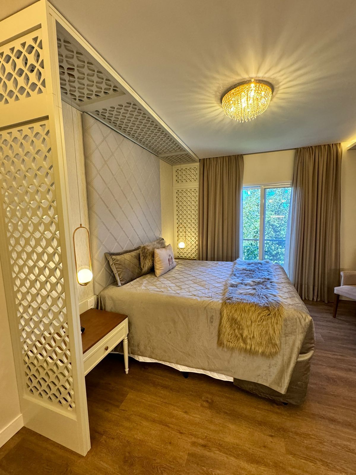 Ein Schlafzimmer mit einem großen Bett, einem Nachttisch, einem Sessel, einer dekorativen Deckenleuchte und einem Fenster mit Vorhängen.
