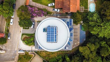 Una veduta aerea di un edificio con tetto circolare, circondato da auto parcheggiate, alberi e una piscina.