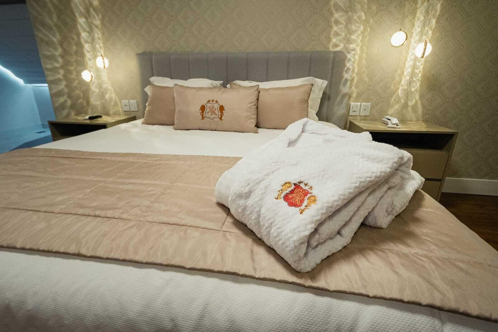 A neatly made bed with a beige bedspread and pillows, a white towel with a crest, and two bedside lamps on a nightstand.
