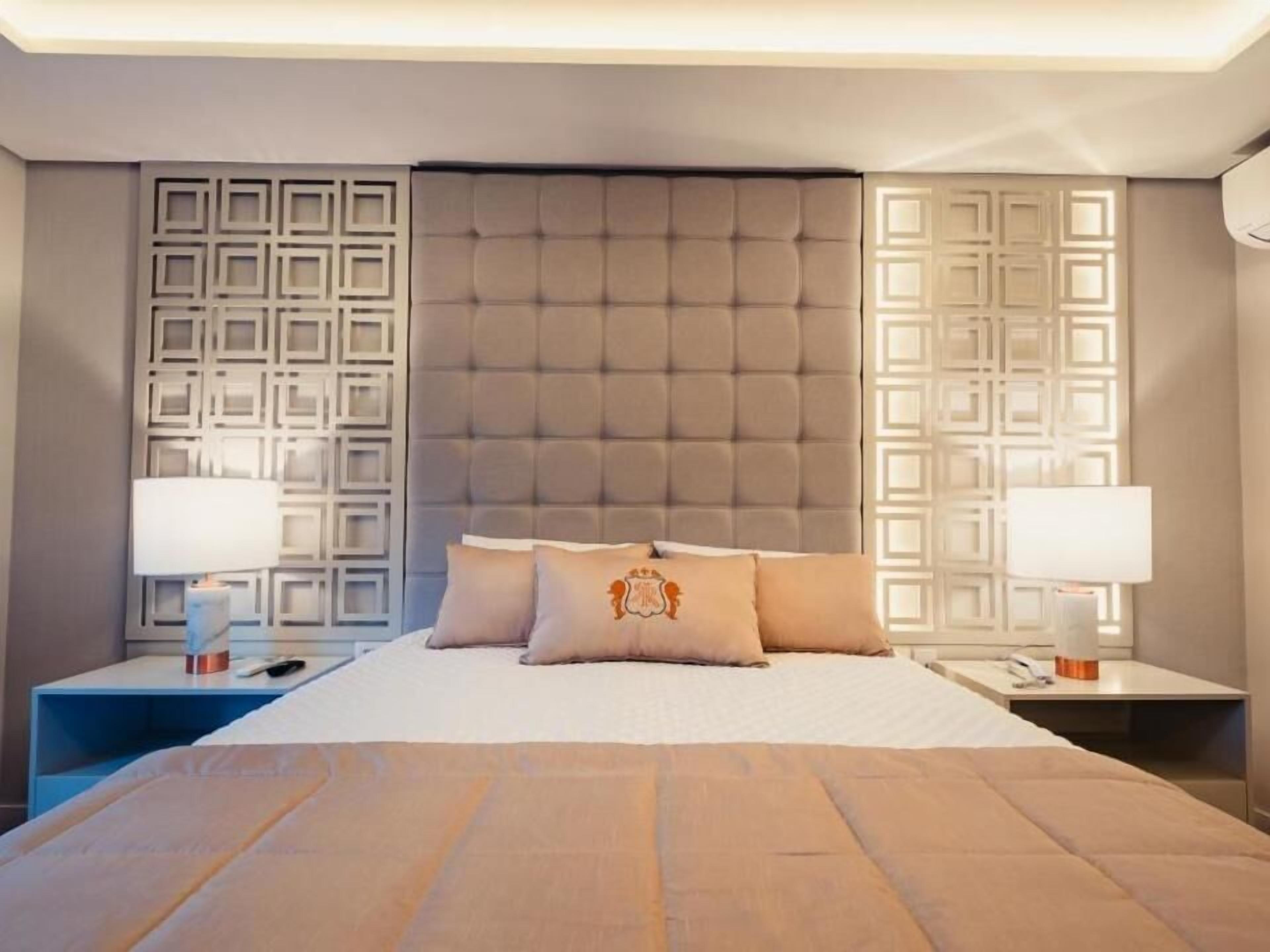 A modern hotel room with a large bed, tufted headboard, decorative wall panels, and bedside lamps.