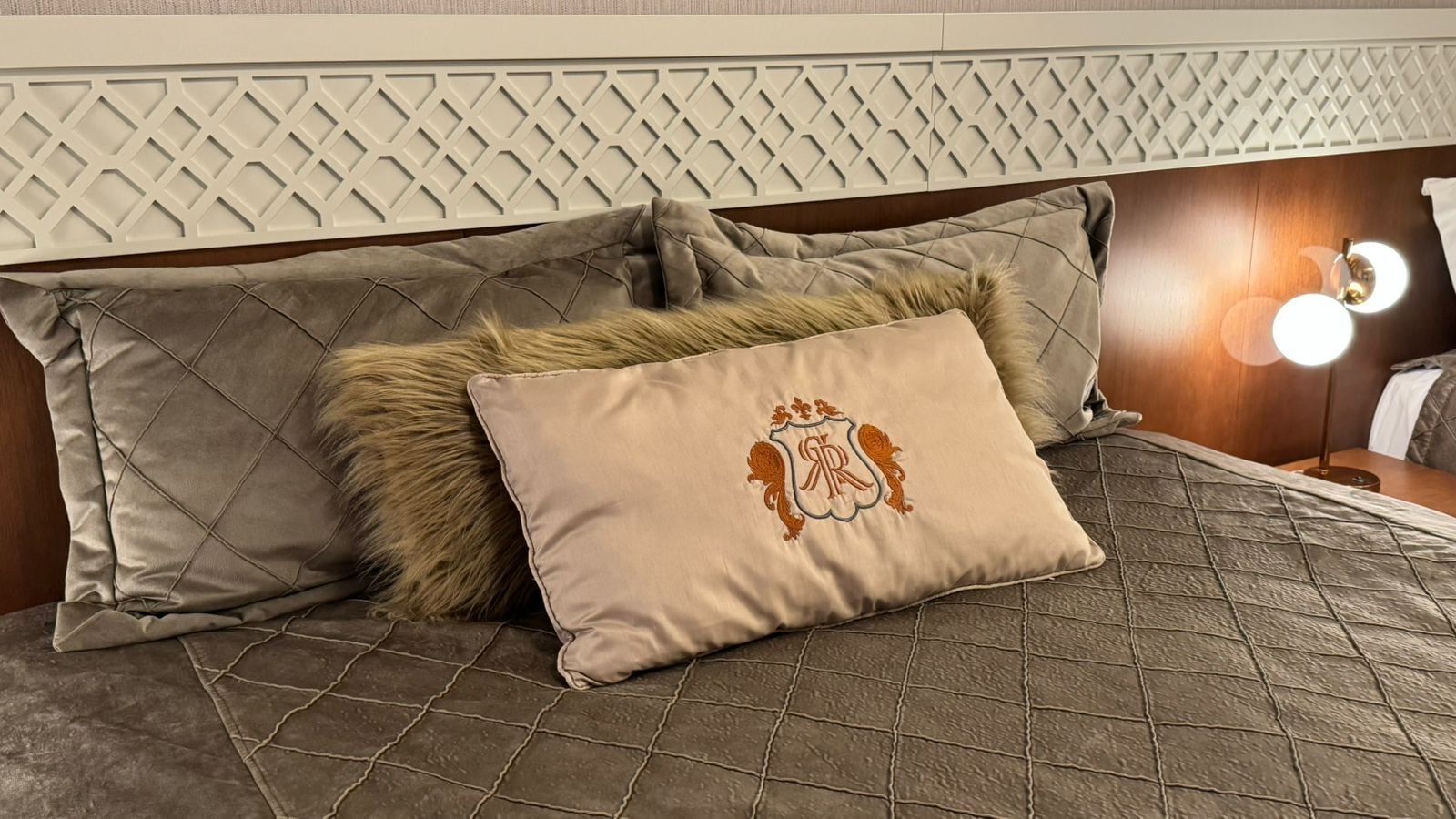 A bed with a quilted cover, two pillows, one with a monogram, and a headboard with a geometric pattern.