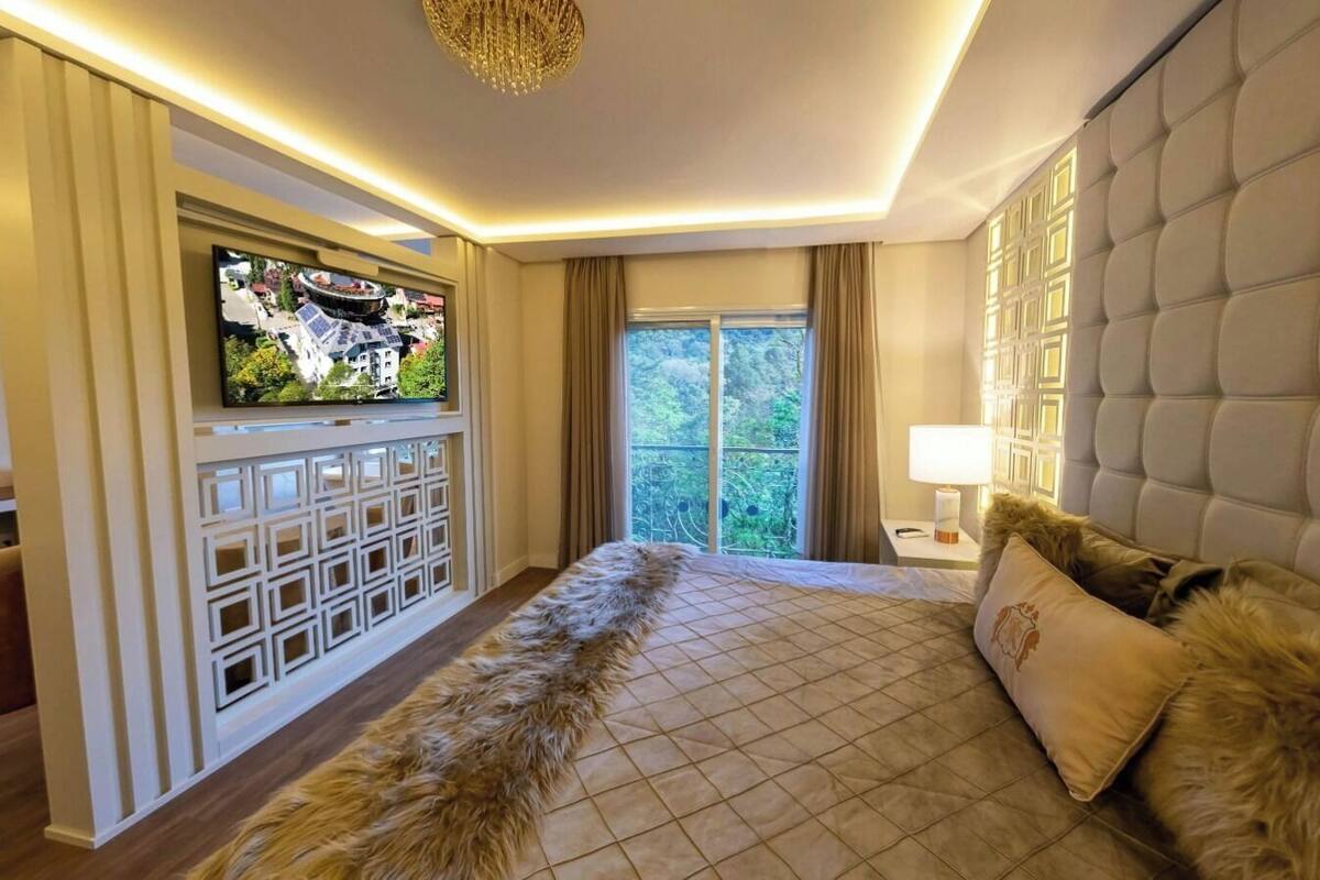 A modern hotel room with a large bed, a television mounted on the wall, and a view of greenery outside.