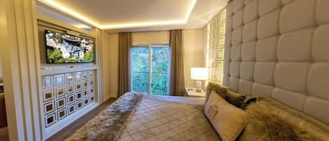 A modern hotel room with a large bed, a television mounted on the wall, and a view of greenery outside.