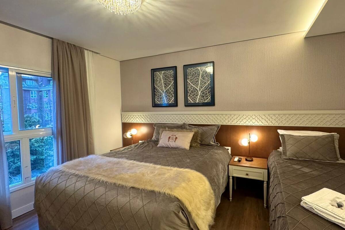 A hotel room with two beds, a large window with curtains, a chandelier, and two framed wall art pieces.