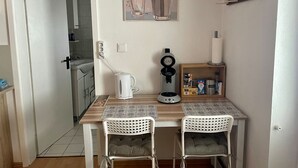 Un petit coin repas avec une table et deux chaises, une machine à café, une bouilloire et une étagère contenant divers produits à base de café.
