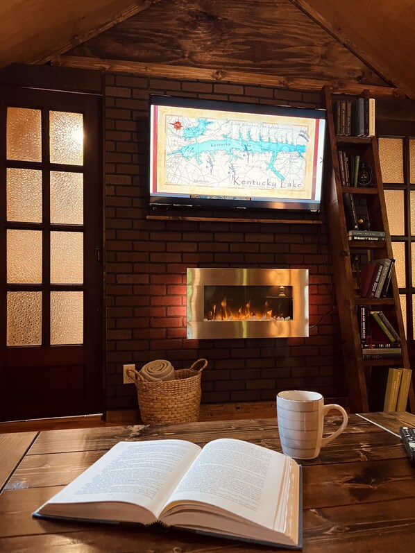 Una habitación acogedora con chimenea, un televisor sobre ella mostrando un mapa, una estantería y un libro abierto sobre una mesa de madera.
