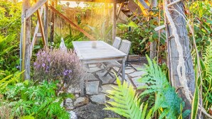 Une terrasse en bois avec une table et des chaises, entourée d’une végétation luxuriante et bordée d’un chemin en pierre.