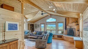 A cozy cabin interior with a wooden ceiling, a fireplace, a large window, and a seating area with a sofa and armchairs.