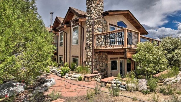 A large, multi-story stone building with a balcony, surrounded by trees and rocks.