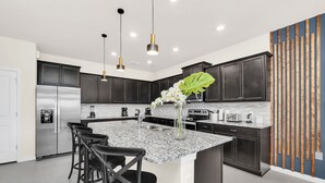 A modern kitchen with dark cabinets, a stainless steel refrigerator, a marble countertop, and a central island with bar stools.