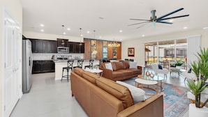 A spacious living area with a brown leather sofa, a coffee table, and a dining table with chairs. There is a kitchen area with dark cabinets and a refrigerator.