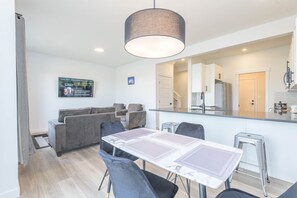 A modern living room with a dining table, grey sofas, and a large mirror reflecting the kitchen area.