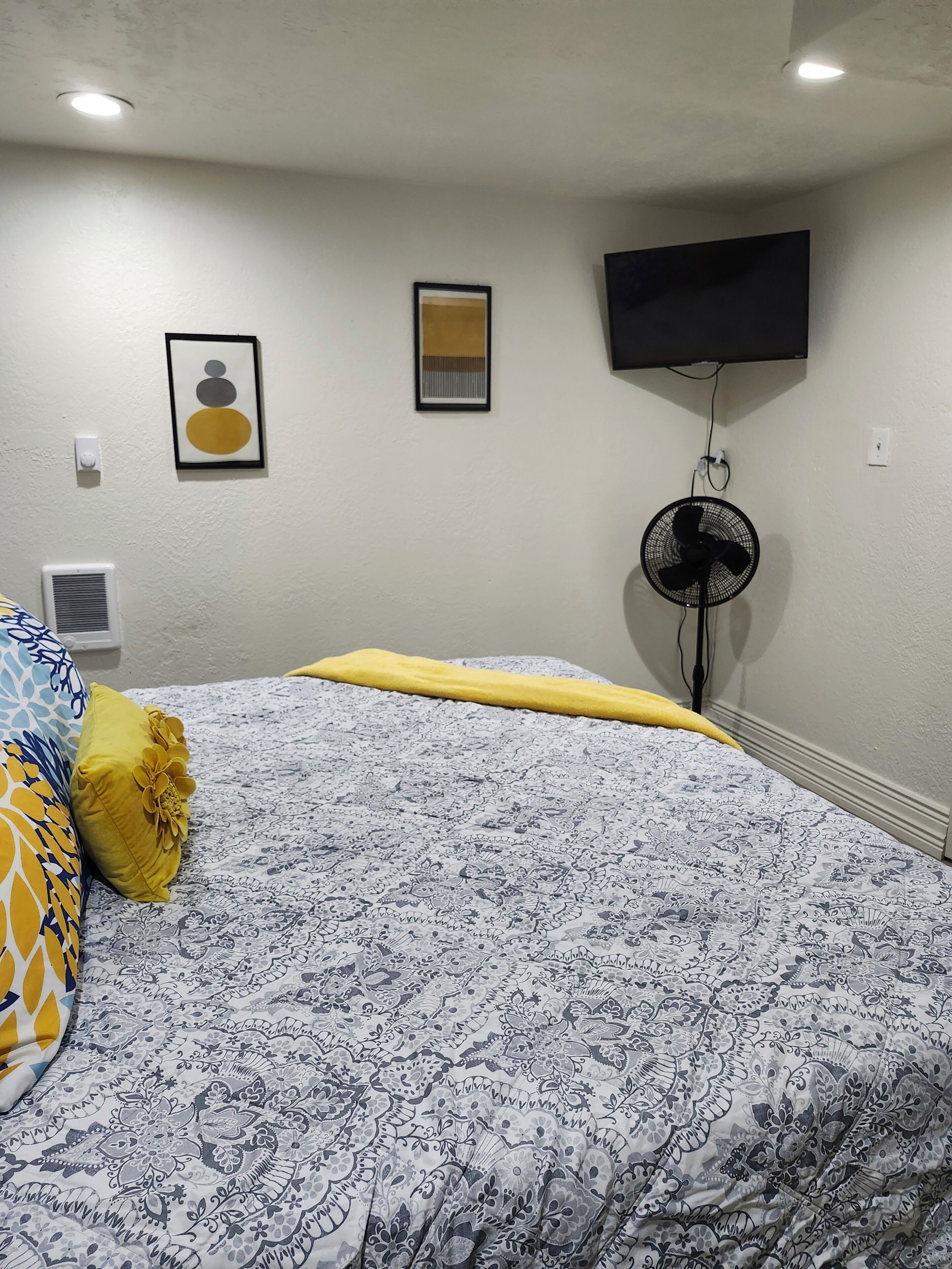 Ein Schlafzimmer mit einem gemusterten Bettbezug, einem flachbildfernseher an der Wand, einem Standventilator und einer Heizungseinheit.