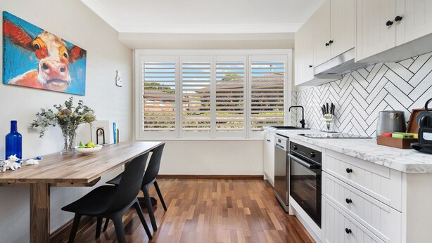 Una cocina moderna con armarios blancos, una mesa de comedor de madera y un ventanal con persianas.