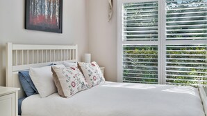Un dormitorio con cama blanca, almohadas estampadas, una mesita de noche, una obra enmarcada en la pared y una ventana con persianas.