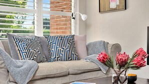 Una sala de estar con un sofá color beige, cojines estampados, un florero de vidrio con flores y una revista sobre una mesa de centro.
