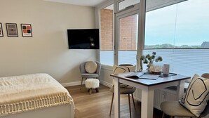 A modern bedroom with a bed, a desk, and a chair. There is a TV mounted on the wall, a window with a view, and a small table with decorative items. - Salzwasser - Modernes App. mit Balkon, Meerblick, Pkw-stellplatz, Wlan. Haustiere Erlaubt (List)