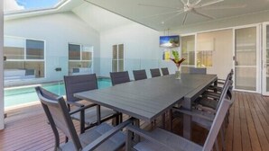 Uma área moderna para refeições ao ar livre, com uma mesa comprida, cadeiras, ventilador de teto e vista para uma piscina.