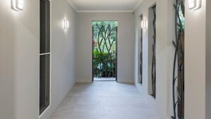 Corredor com paredes brancas, iluminação embutida e vista para o verde através de uma janela.