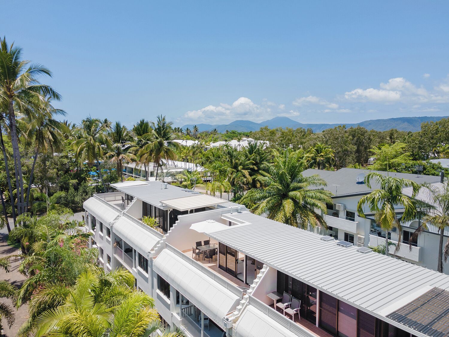 A modern multi-story hotel with a rooftop terrace, surrounded by palm trees and a mountainous backdrop.
