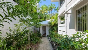 A narrow pathway flanked by lush greenery and white buildings, leading to a door. - Apartment 2 @ Ocean Palms Apartments (Douglas)