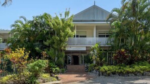 A two-story building with a balcony, surrounded by tropical plants and a gravel pathway leading to the entrance. - Apartment 2 @ Ocean Palms Apartments (Douglas)