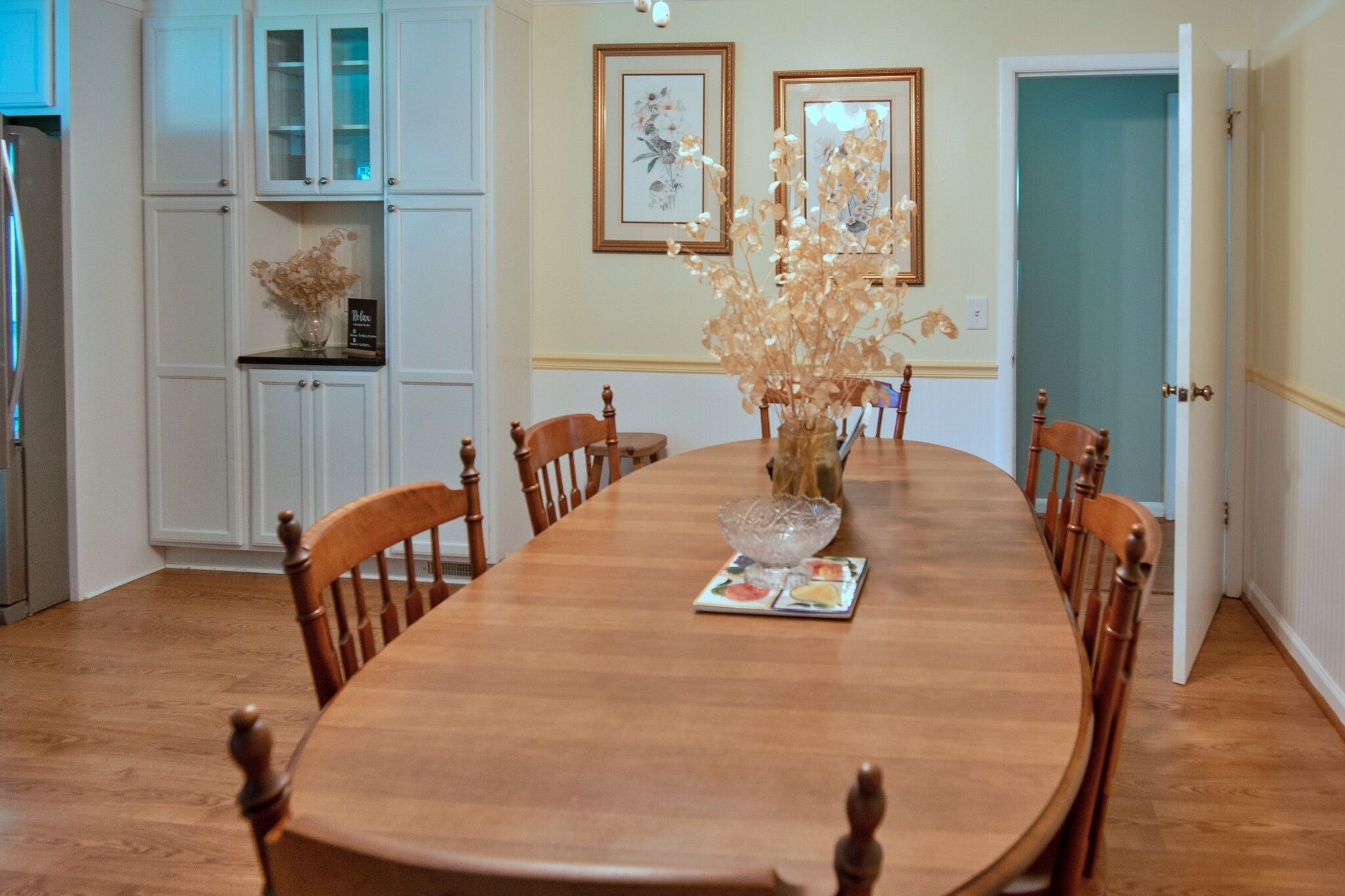 Une salle à manger avec une table en bois, des chaises et un vase contenant des fleurs séchées. On aperçoit des meubles blancs et une porte au fond.