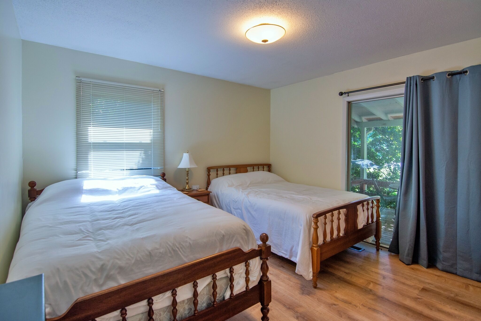 A bedroom with two wooden beds, a nightstand with a lamp, a window with blinds, and a view of greenery outside.