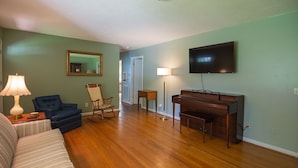 A living room with a sofa, a blue armchair, a wooden piano, a television, and a lamp.